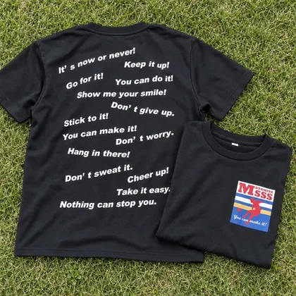 黒いTシャツの背中いっぱいに英語の応援の言葉を白で。左胸にフルカラーのエンブレム